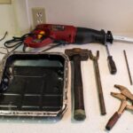 Close-up of a pickle fork tool holding a potato, alongside other car parts tools like a trim removal tool and vice grips, set against a backdrop of automotive parts.