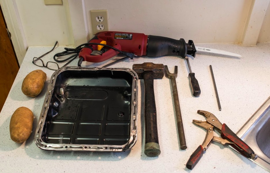 Close-up of a pickle fork tool holding a potato, alongside other car parts tools like a trim removal tool and vice grips, set against a backdrop of automotive parts.